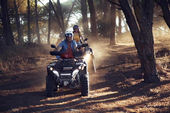 Two people riding all terrain vehicles, or ATVs, through a sunkissed forest