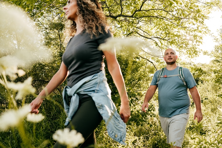 Two people looking away from the camera and walking in hiking clothes through trees and flowers that are close to the camera