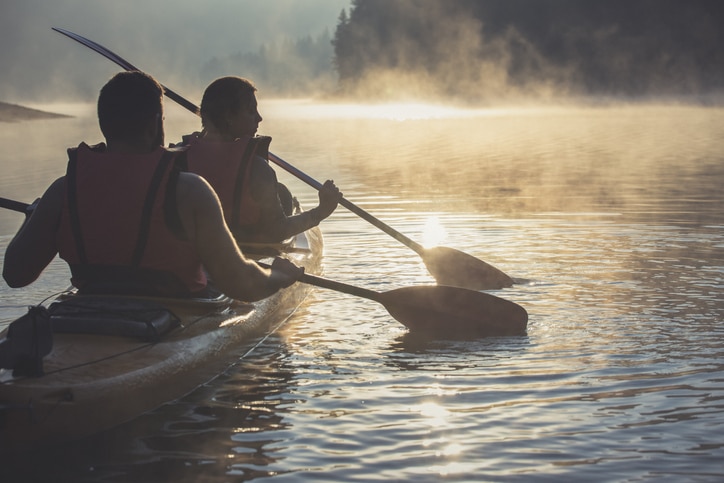 Two people riding a two-manned kayak together, with both of their paddles sitting in the water on the right hand side of the boat.