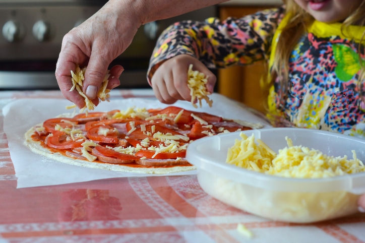 An adult and a child making a home made pizza, holding cheese over the pizza