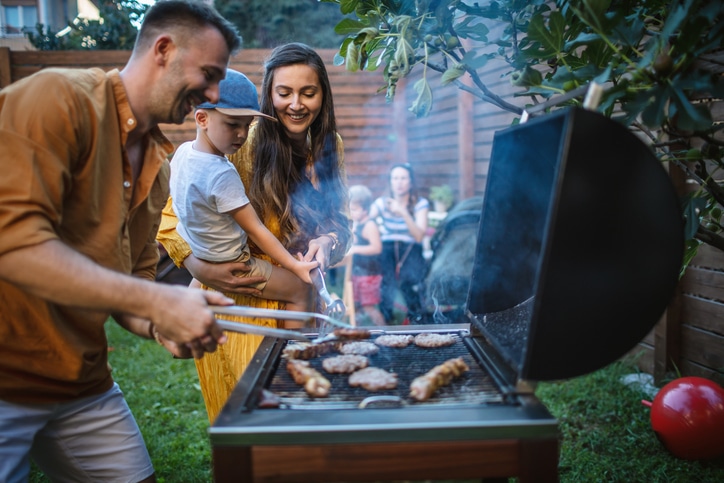 A man in a brown shirt holds metal tongs over a grill with meats, assisted by a woman holding a toddler as they watch and smile