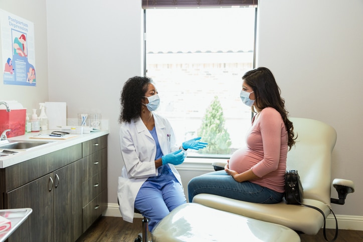 A pregnant woman sitting with a medical provider wearing a mask and gesturing with her hands, used to explain prenatal care
