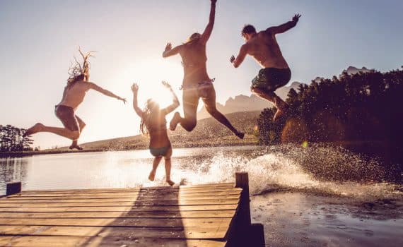 A group of four people jumping off a dock into a natural body of water, used to explain activities during the summer