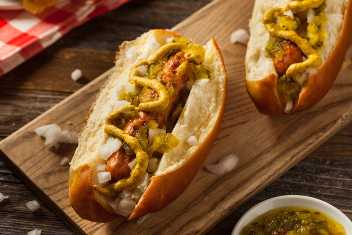 A photograph of two sausage bun with mustard, onions, relish on a wooden board