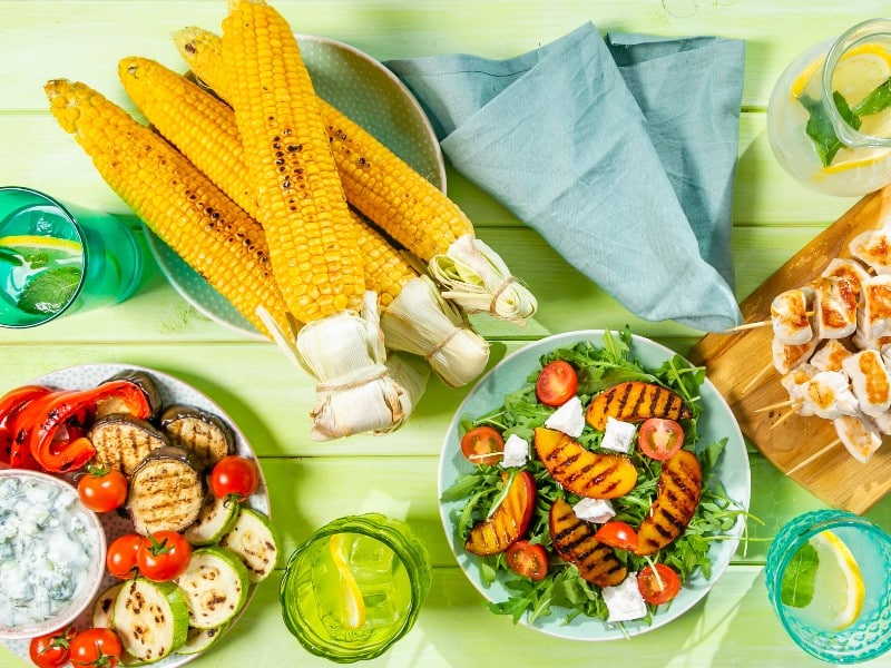 An array of healthy summer food, including grilled fruit salad, grilled corn on the cobb, and grilled squash and zucchini