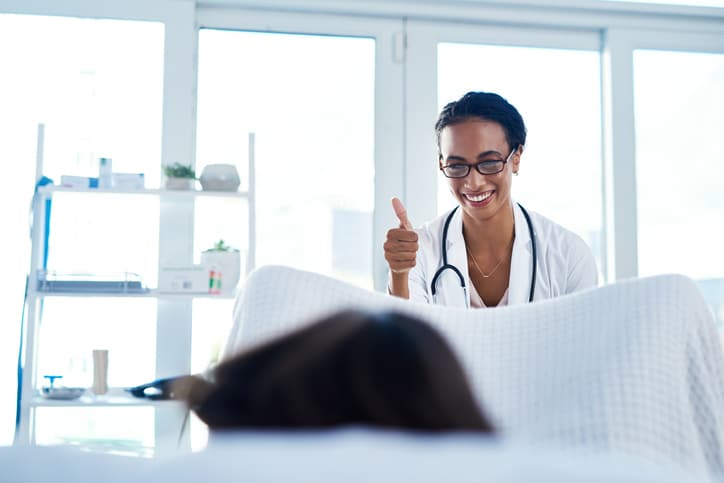A stock image of a women's health physician giving the thumbs up toward a person in the foreground looking away from the camera lying on a medical examination table