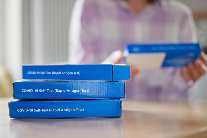 A stack of three COVID-19 home test kits stacked on a table in the foreground with someone reading a different box in the background