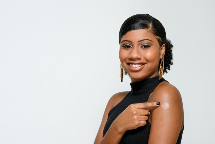 Stock image of a black woman smiling and looking at the camera, pointing to a round bandage on her upper arm