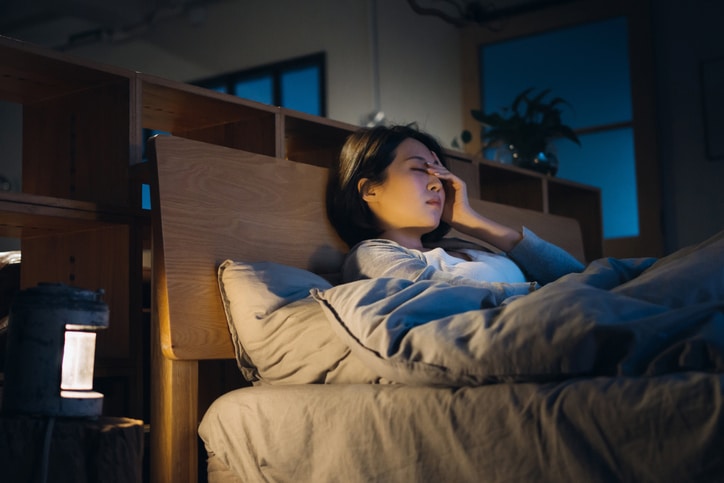 A stock photograph of a woman in bed, sitting against the headboard with her eyes closed, holding her forehead with her fingertips