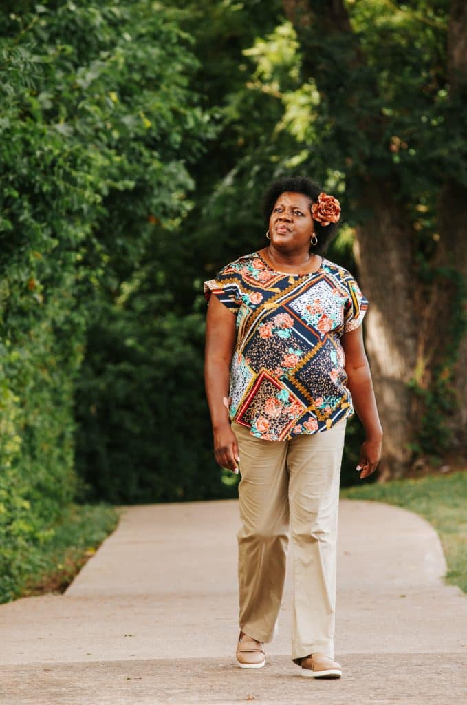 Renee Cooper wearing brown slacks, a colorful floral top, and a flower clip in her hair, photographed outside