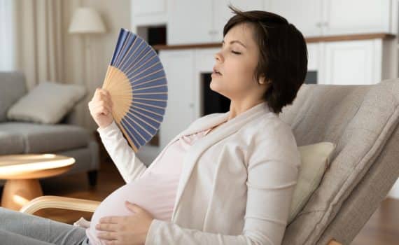 Pregnant woman resting her left hand on her abdomen while using a handheld blue fan with her right hand