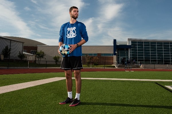 Zach Thibodeau holding a blue, black, and yellow soccer ball while standing on a soccer field and looking off to the side of the frame