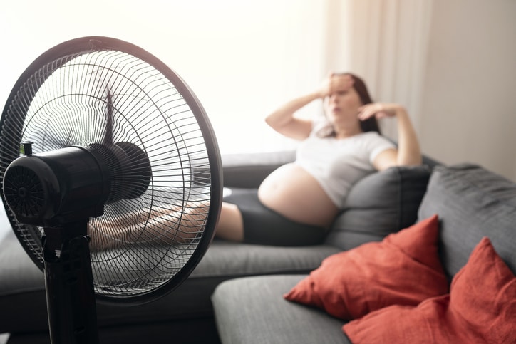 Pregnant woman in background holding her forehead and fanning here face, with a black standing fan in the foreground