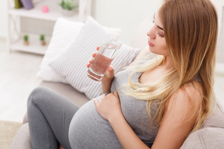 A pregnant woman in gray pants and a gray tank top rests her hand on her stomach while holding a class of water