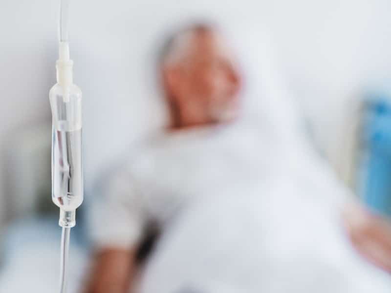 A stock photograph with the foreground focused on an IV and the background a blurred image of a man in a hospital bed, used to explain a new prostate cancer drug