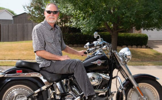 David Fast sitting on a Harley Davidson motorcycle while wearing a gray button down shirt and sunglasses
