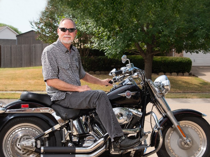 David Fast sitting on a Harley Davidson motorcycle while wearing a gray button down shirt and sunglasses