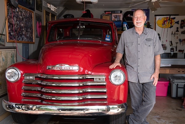 David Fast photographed looking and the camera and smiling with his right hand resting on the front of a bright red vintage Chevrolet pickup truck