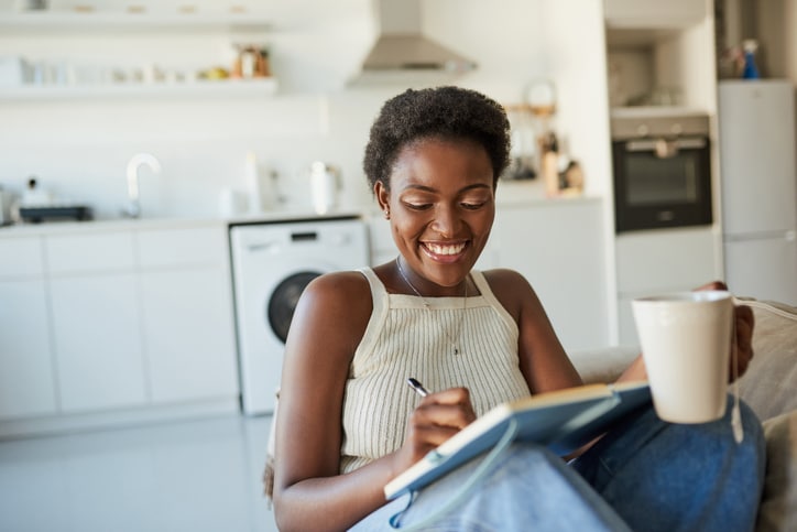 Woman smiling and writing in a notebook while holding a white mug with a tea bag hanging off of it