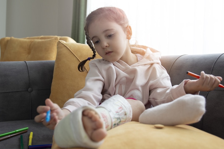 A child holding markers looks down on her leg, which appears to be a in hard cast