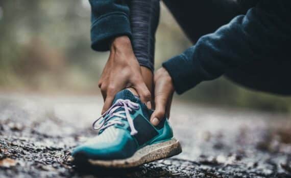 A person who appears to be on a running or hiking trail dressed in bright teal sneakers clutches their ankle with both hands, used to explain strains and broken bones