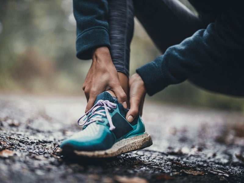 A person who appears to be on a running or hiking trail dressed in bright teal sneakers clutches their ankle with both hands, used to explain strains and broken bones