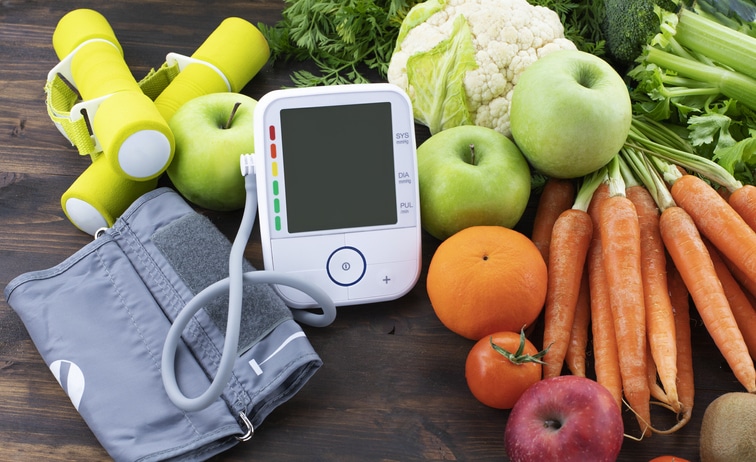 A mobile blood pressure monitor staged in a photograph with healthy foods including carrots, green apples, oranges and more