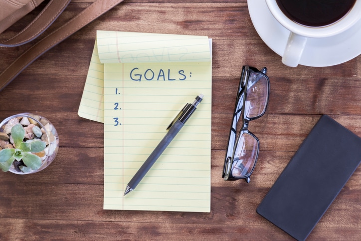 A small notepad with a mechanical pencil resting on top of it with glasses and other items surrounding it, with "Goals" written at the top