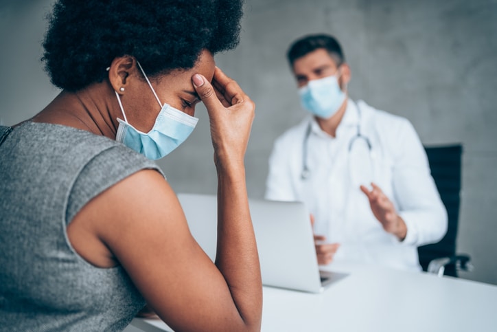 A woman holding her forehead and looking down while a medical professional faces her and looks to be explaining something and talking with his hands.