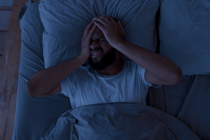 A man lying in bed in a dark room, clutching his head as if he has a headache