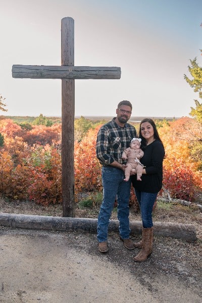 Kaydee Endrody, husband Andy, and baby Ruby all looking at the camera in front of a wooden cross