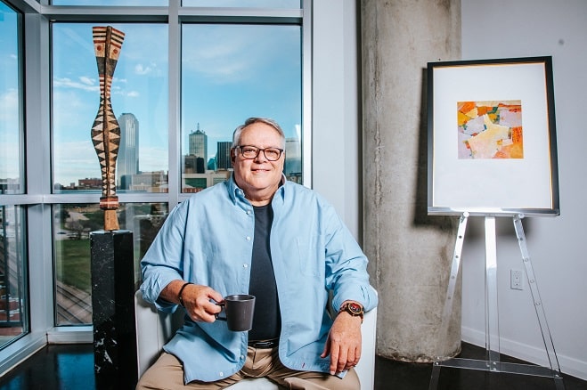 Kerry Walton photographed in blue button down shirt and smiling at the camera in front of a large window, a black framed painting, a piece of wooden art, and holding a mug.