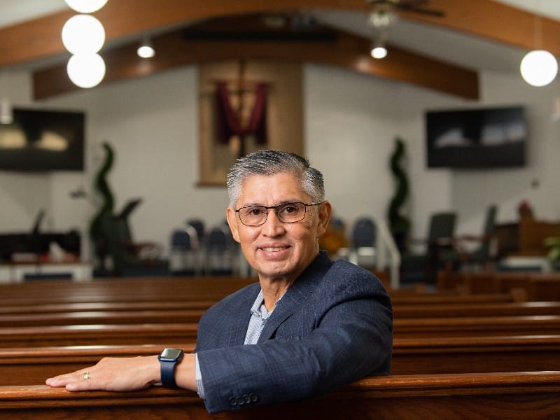 Pastor Arturo Malacara sitting in a church pew looking at the camera, used to explain his widow maker heart attack recovery