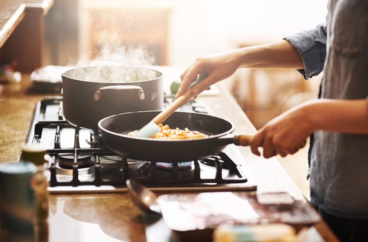 A person cooking over the stovetop with a steaming pot and a pan with colorful veggie that is being stirred 