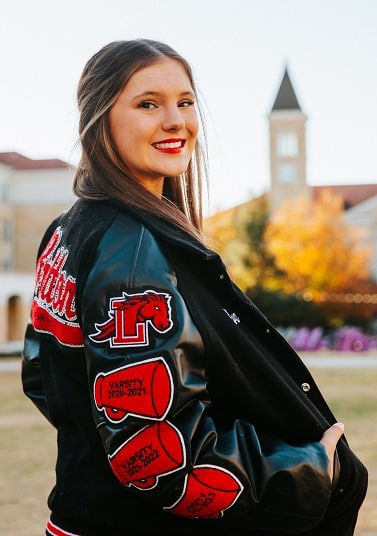 A young woman named Ava Britton wearing a cheerleading uniform and jacket and smiling at the camera 