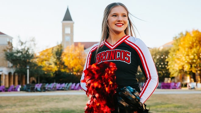 A young woman named Ava Britton wearing a cheerleading uniform and smiling at the camera and holding two pom poms, black and red, close to the camera