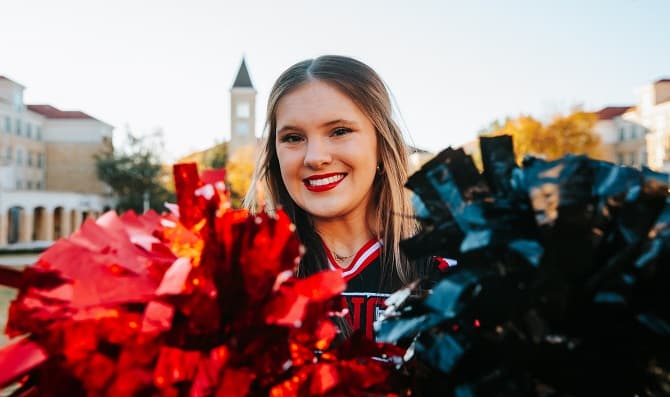 A young woman named Ava Britton wearing a cheerleading uniform and smiling at the camera and holding two pom poms, black and red, close to the camera