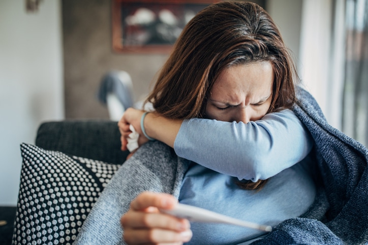 A woman coughing into her elbow and holding a thermometer. 