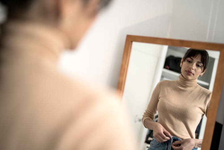 Woman looks at herself in a mirror while zipping up her jeans