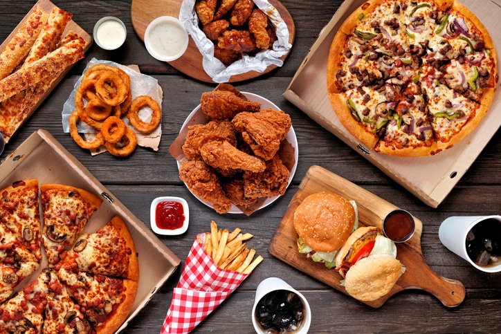 An array of foods laid out on a table as a spread, including onion rings, fried chicken, boneless chicken, pizza, burgers, garlic cheese sticks, french fries, cola, and dipping sauces