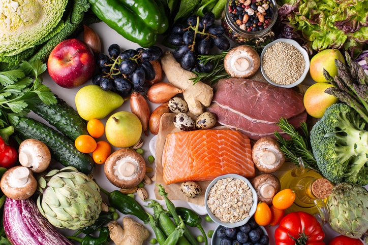 An array of healthy foods as a spread, including mushrooms, eggplant, artichoke, salmon, pears, shallots, steak, oats, quail eggs, broccoli, oil, tomatoes, lettuce, beans, grapes, peppers, herbs, cucumbers, blueberries, ginger, seeds, and moer.