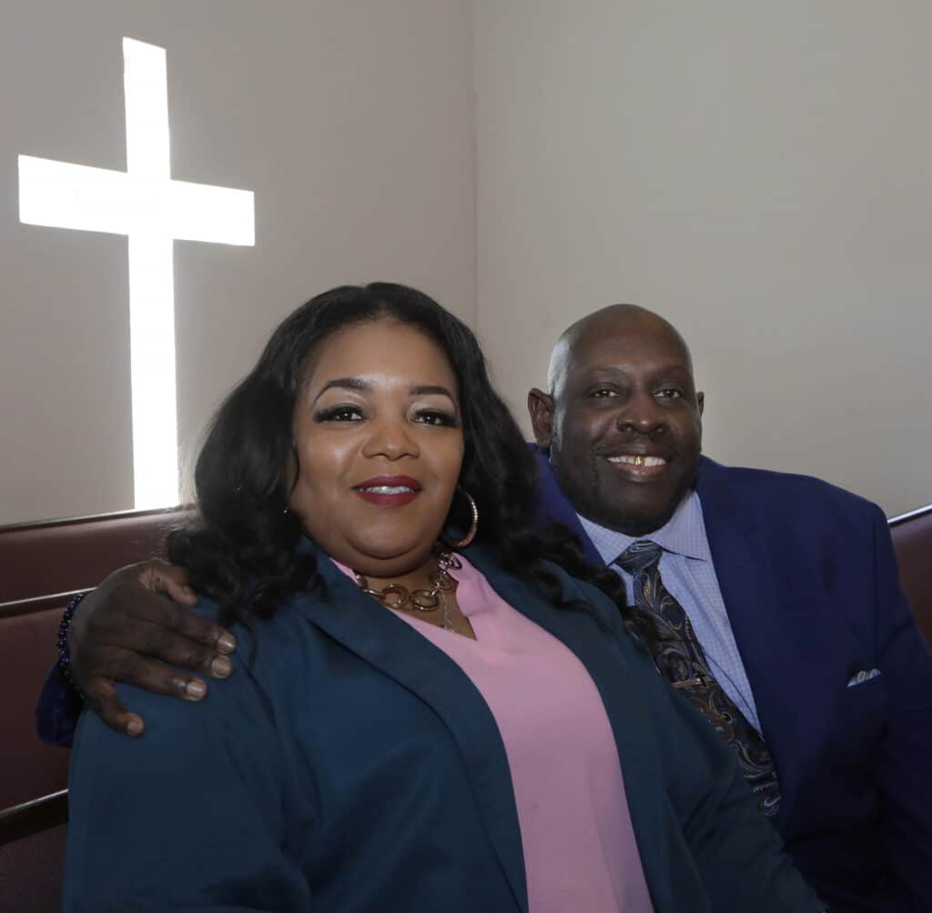 Pastor Leonard Johnson with his arm around his wife, Tammy, photographed in a church with a cross projected on the wall behind them.