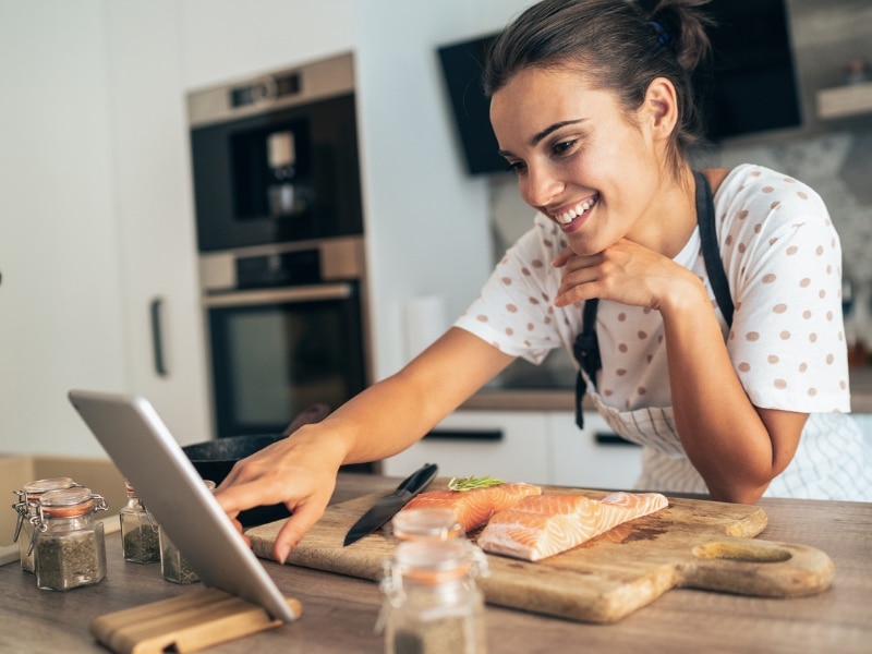 Woman looks at tablet smiling and reading along while in the kitchen preparing some salmon to cook, used to describe the relationship between certain foods and fertility