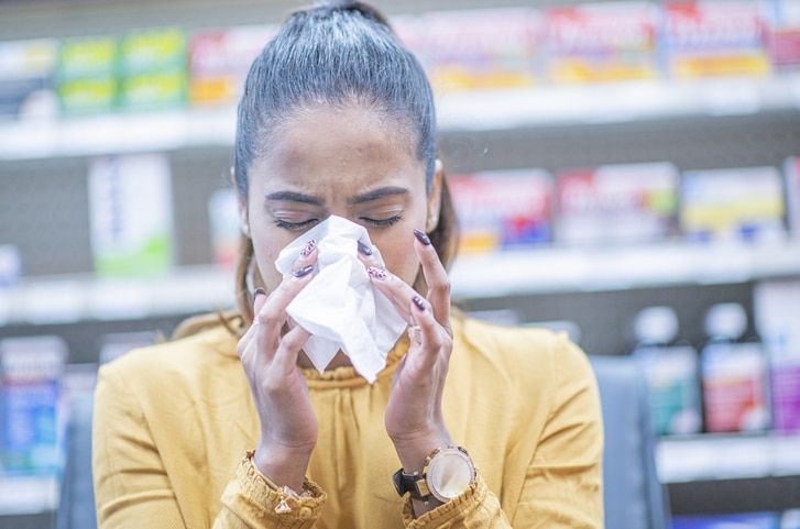 A young woman stands in a store in front of a shelf containing antihistamines. She is blowing her nose.