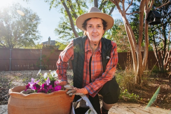 Margaret Adams smiling at the camera for a photo, standing outside in gardening attire next to a pot of flowers