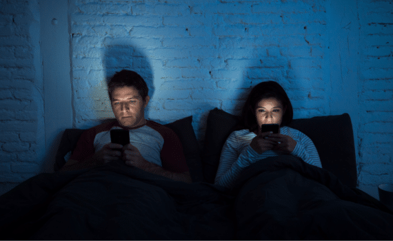 Two people sitting in bed in a dark room, illuminated by the cell phones in front of their faces