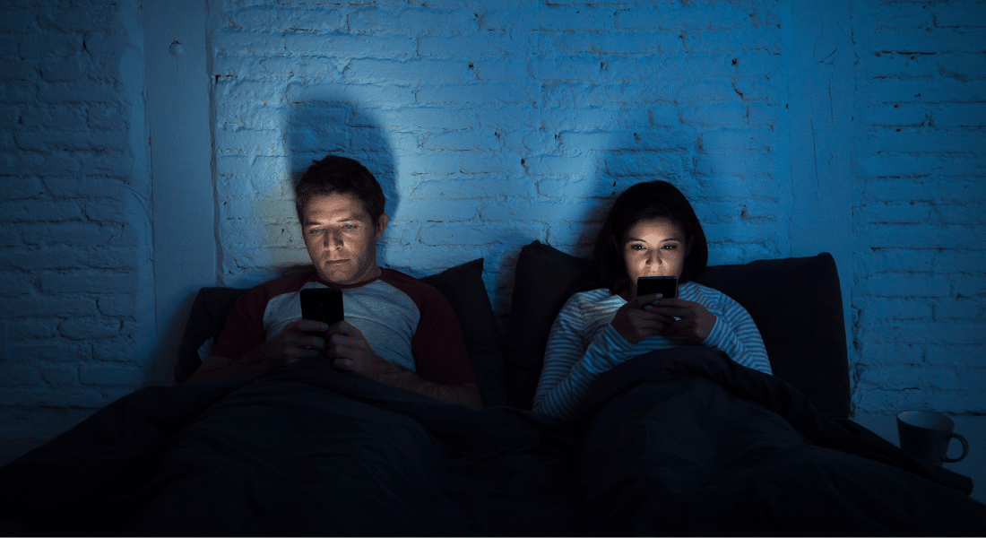 Two people sitting in bed in a dark room, illuminated by the cell phones in front of their faces