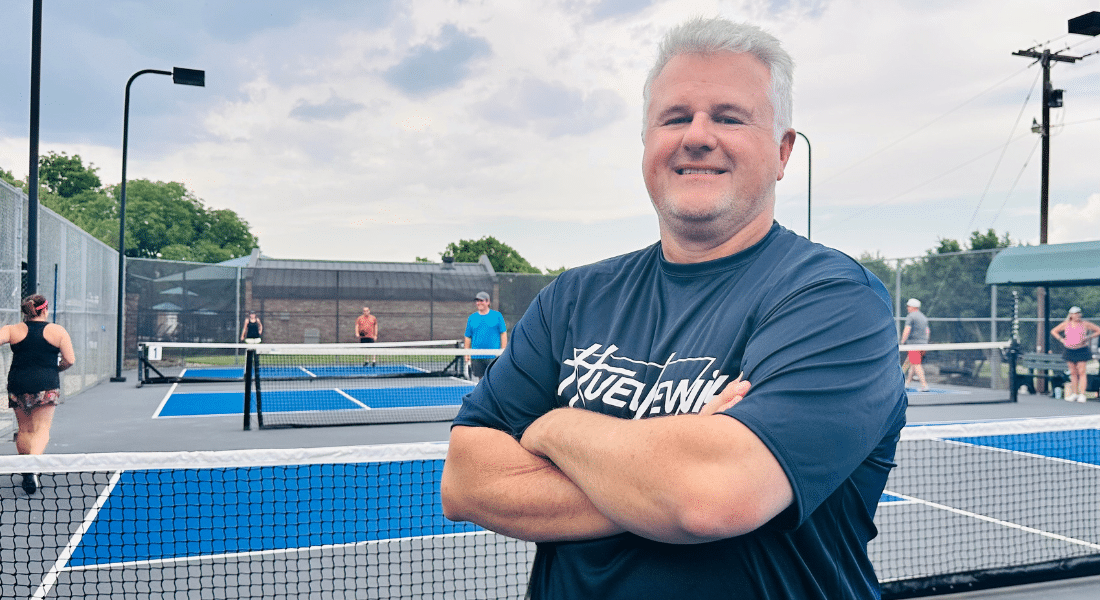 Gary Davis, resident of the Dallas Fort Worth area, photographed smiling with his arms crossed in front of a pickleball court, used to explain his recovery from blood clots in the lungs