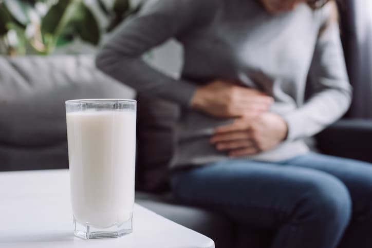 A glass of milk stands on a table, while in the background a woman holds her stomach.