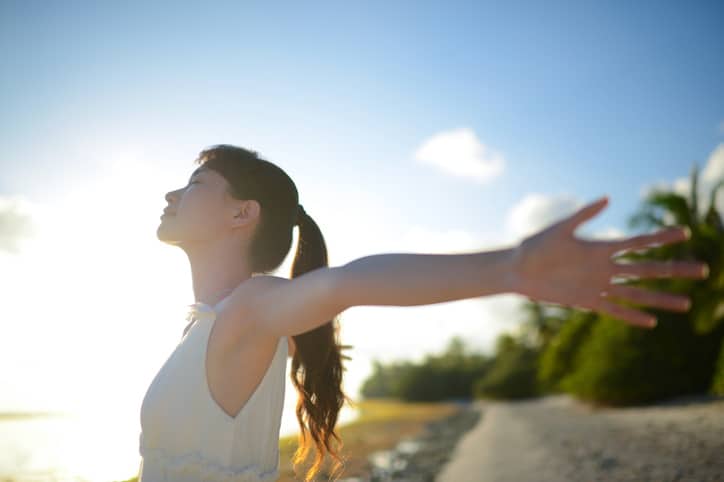 A woman with a white tank top and ponytail standing outside with her arms outstretched